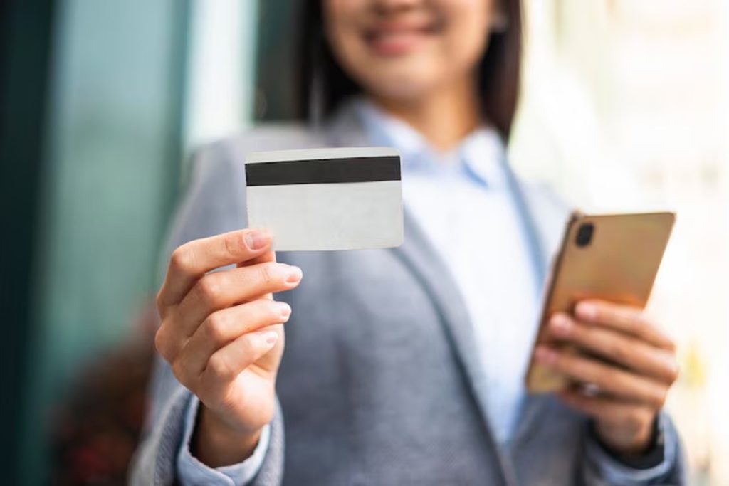 Smiling businesswoman in a blazer holding a smartphone and displaying the back of a blank credit card — a professional visual representing credit cards for small businesses.