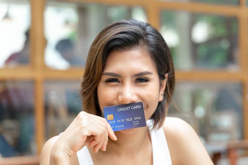 Smiling young woman holding a credit card close to her face in a relaxed café setting — representing the ease and convenience of using credit cards with zero international fees.
