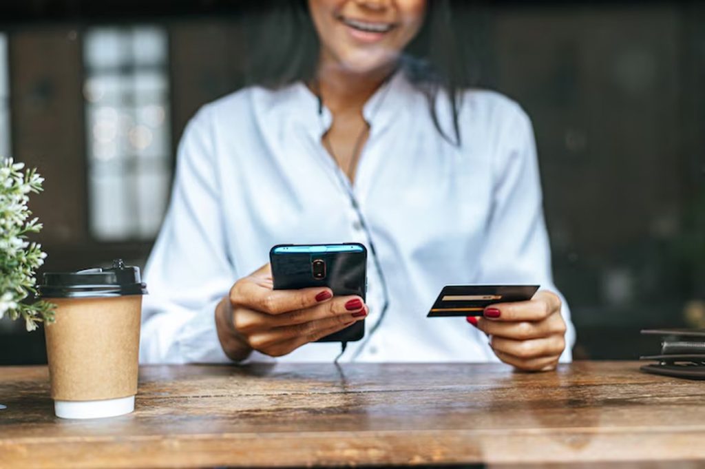 Woman sitting at a café table, smiling while holding a smartphone and a credit card — a casual scene that reflects the everyday convenience and benefits of credit cards in modern life.