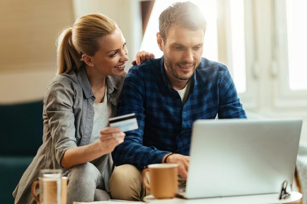Smiling couple sitting on a couch with a laptop and credit card, making an online purchase together — a relaxed moment that highlights the role of credit cards on the financial planning for modern households.