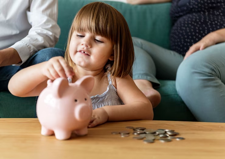 A young girl placing coins into a pink piggy bank while sitting at a table with her family in the background—representing the early development of positive financial habits and the importance of teaching children how to save money.