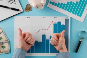 A pair of hands giving thumbs up in front of a financial growth chart with bars and a rising red trend line, surrounded by coins, dollar bills, a piggy bank, and graphs—symbolizing positive outcomes from setting clear financial priorities.