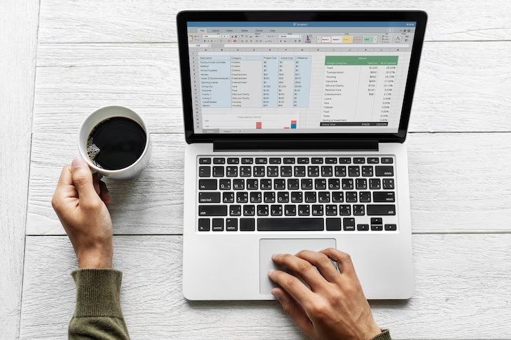 A person working on a laptop while holding a cup of coffee, reviewing a spreadsheet with charts and financial data—demonstrating the use of organized lists to control monthly expenses effectively.