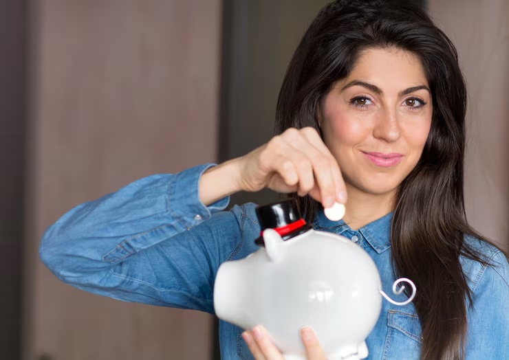 A smiling woman in a denim shirt placing a coin into a white piggy bank, symbolizing the importance of taking steps to organize financially and build personal savings.