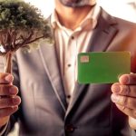 Homem de terno segurando um cartão verde em uma mão e uma pequena árvore na outra, simbolizando cartões sustentáveis e responsabilidade ambiental.