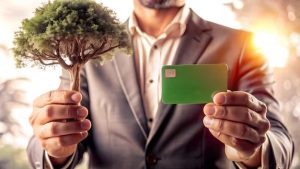 Homem de terno segurando um cartão verde em uma mão e uma pequena árvore na outra, simbolizando cartões sustentáveis e responsabilidade ambiental.