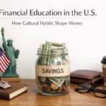 A clean desktop scene representing financial education in the United States, featuring a glass jar labeled “Savings” filled with U.S. dollar bills and coins. On the table are a small Statue of Liberty figurine, an American flag, a leather wallet, a notebook with keys on top, eyeglasses, and a takeaway coffee cup, symbolizing savings habits, personal finance, and American cultural influences on money management.